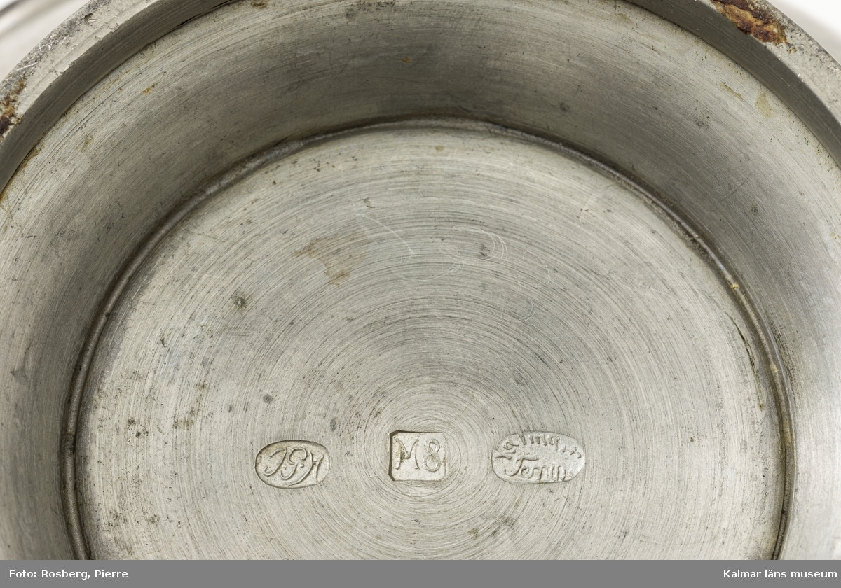 KLM 46718. Vas. Av tenn. Klotformad vas med handtag och fot. På ena sidan en rund platta med motiv i relief föreställande Kalmar slott. På undersidan stämplar: JGH, M8 samt KalmarTenn.