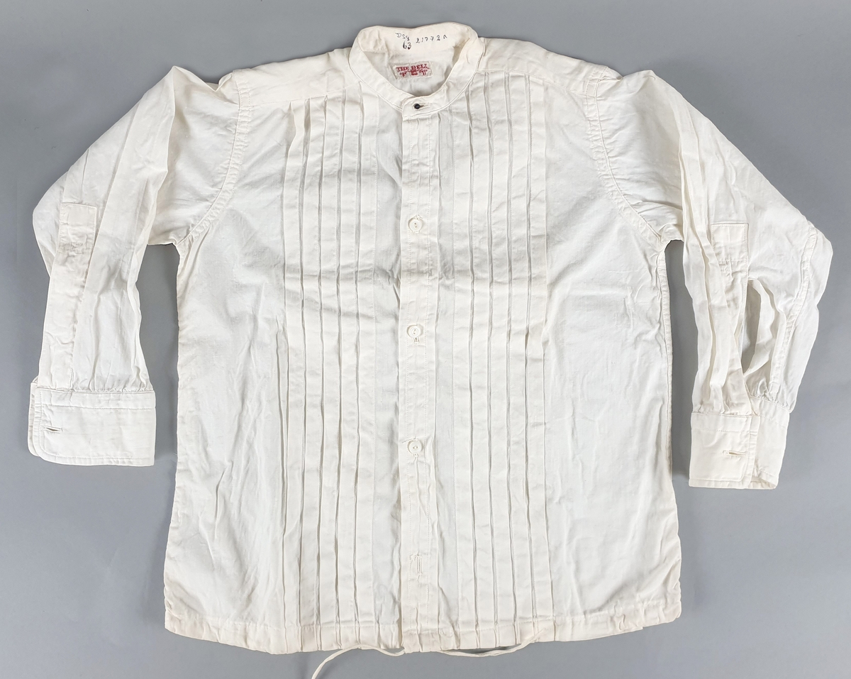 Skjorte av bomull, med vertikale dekorfolder på brystet, snor nederst. Smal linning i halsen med svart mansjettknapp. Hvite knapper midt foran. Skjortene er stivet.