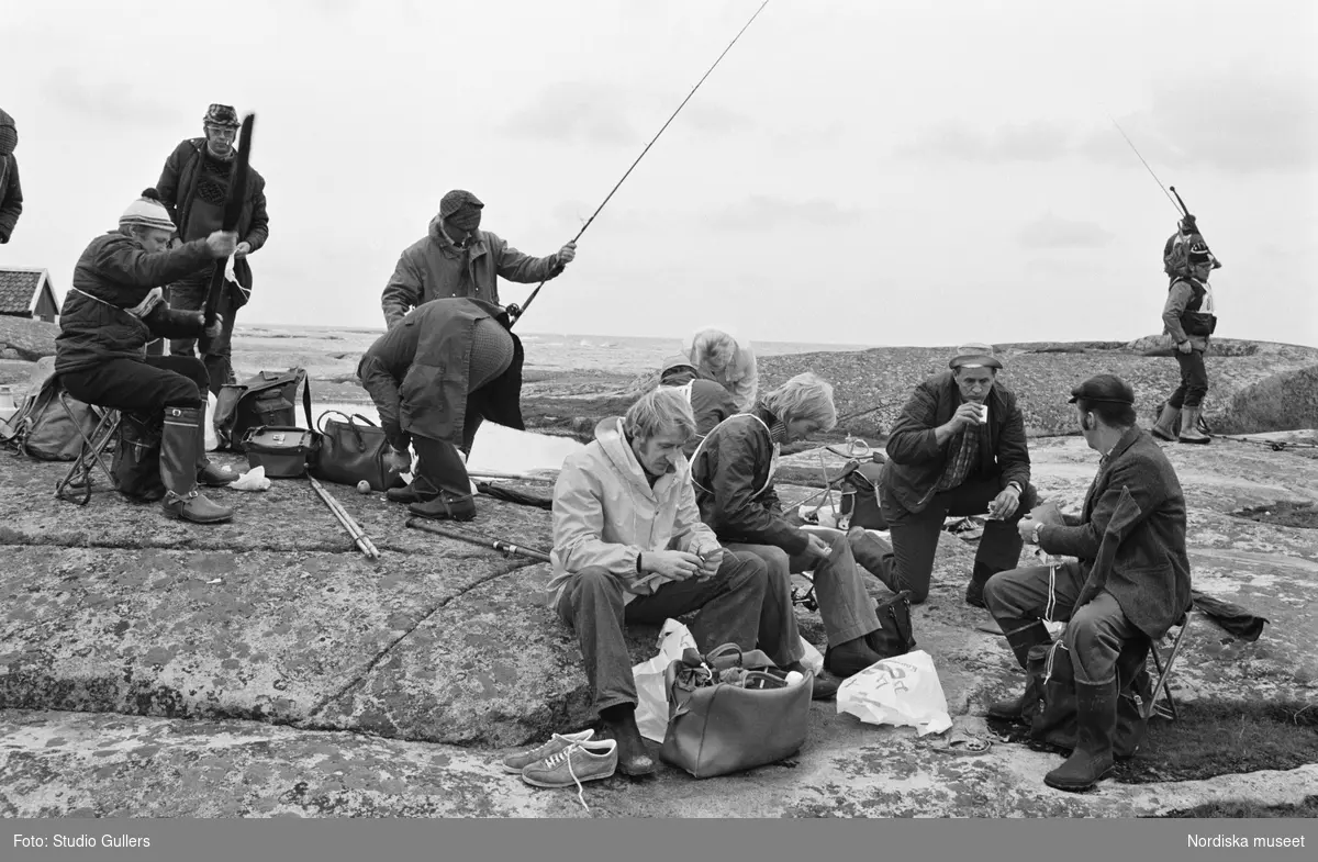 SM i Kustfiske 1976. Några män sitter i förgrunden och dricker kaffe, medan de i bakgrunden är i färd med fisket.
