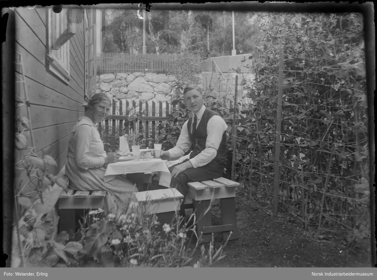 Mann og kvinne sittende ved bord i hage inntil bolighus i Rødbyen. Steinmur på andre siden av stakittgjerde. Vekster bundet opp med trå bak mannen. Trapp opp fra Birkelandsgate til Sam Eydesgate i bakgrunnen.