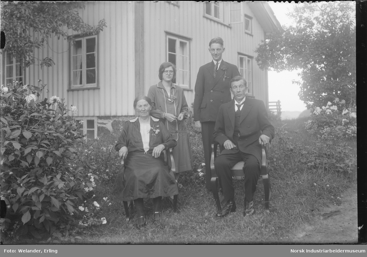 Fire mennesker oppstilt utenfor bolighus. Mann og kvinne sittende i stoler foran to yngre mennesker stående bak. Sannsynligvis ektepar og to barn.