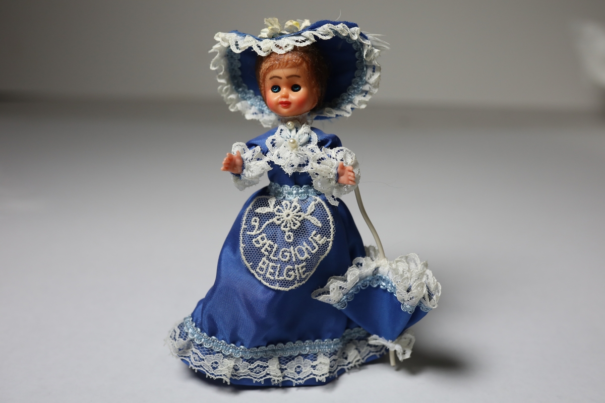 Dukke fra Belgia, stående, med blå kjole i kunstsilke med hvite blondekanter, liknende hatt og parasoll. Har et forkle rundt livet med tekst.