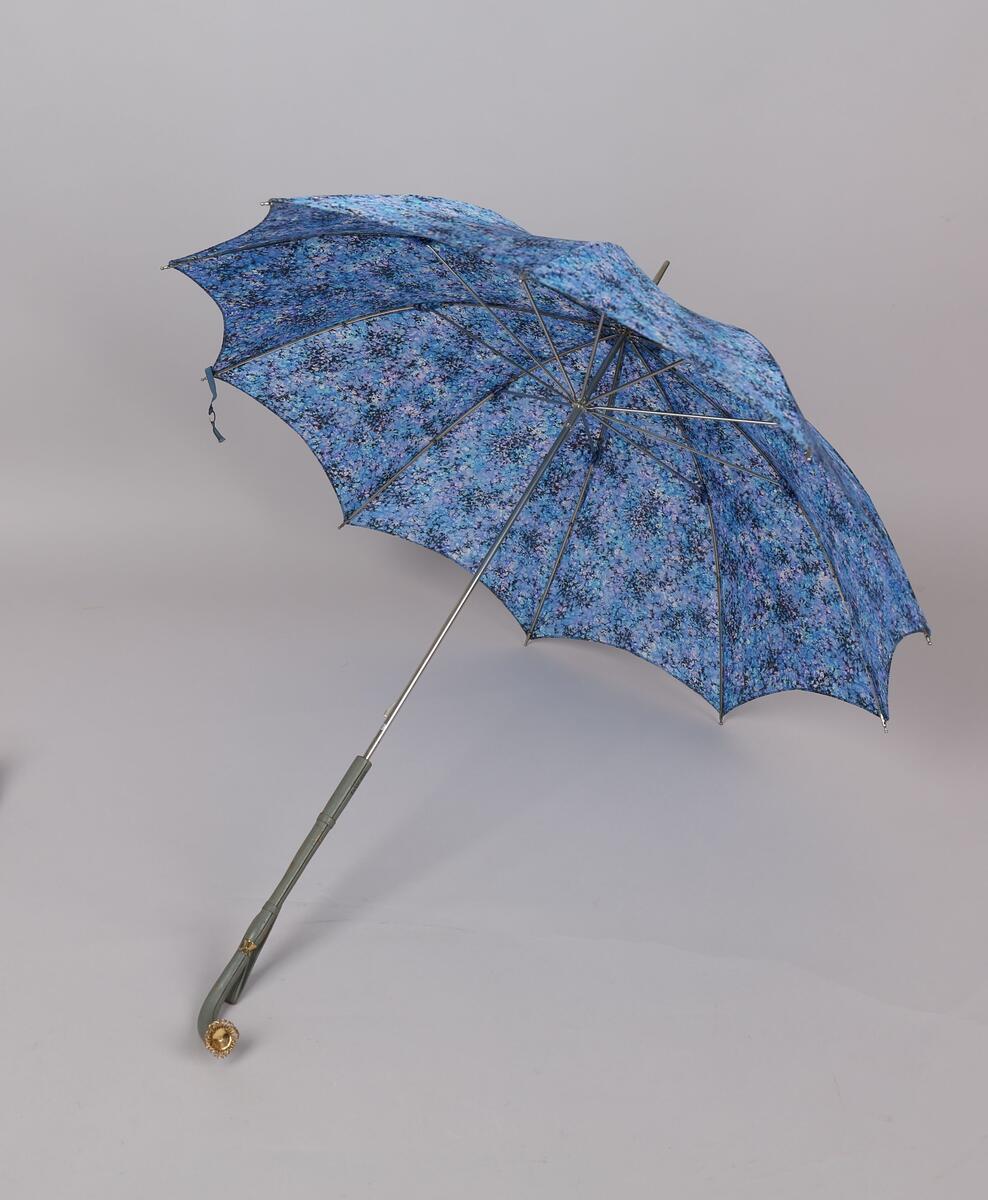 Paraply med 10 spiler og stang i metall.Handtak i skai med dekor i plast, hempe for å ha handa i er festa på handtaket. Duk i kunstfiber i fargane lilla, blå, svart og lys blå, spetta.