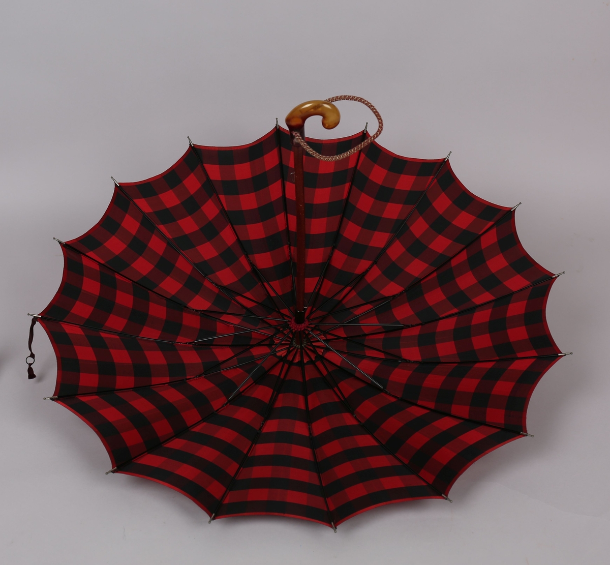 Paraply med 16 metallspilar. Stong i tre. Trekt med stoff i kunstfiber, rutemønster. Handtak i tre med hempe i tekstil til å ta inn på handa. Hempe for lukking av paraplyen.