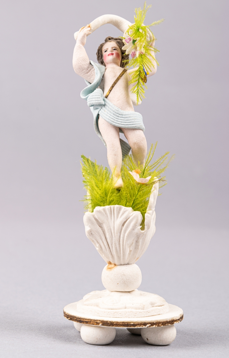 Bröllopskaramell, föreställande ängelfigur/gosse, som står med ymnighetshorn i öppna växtblad.