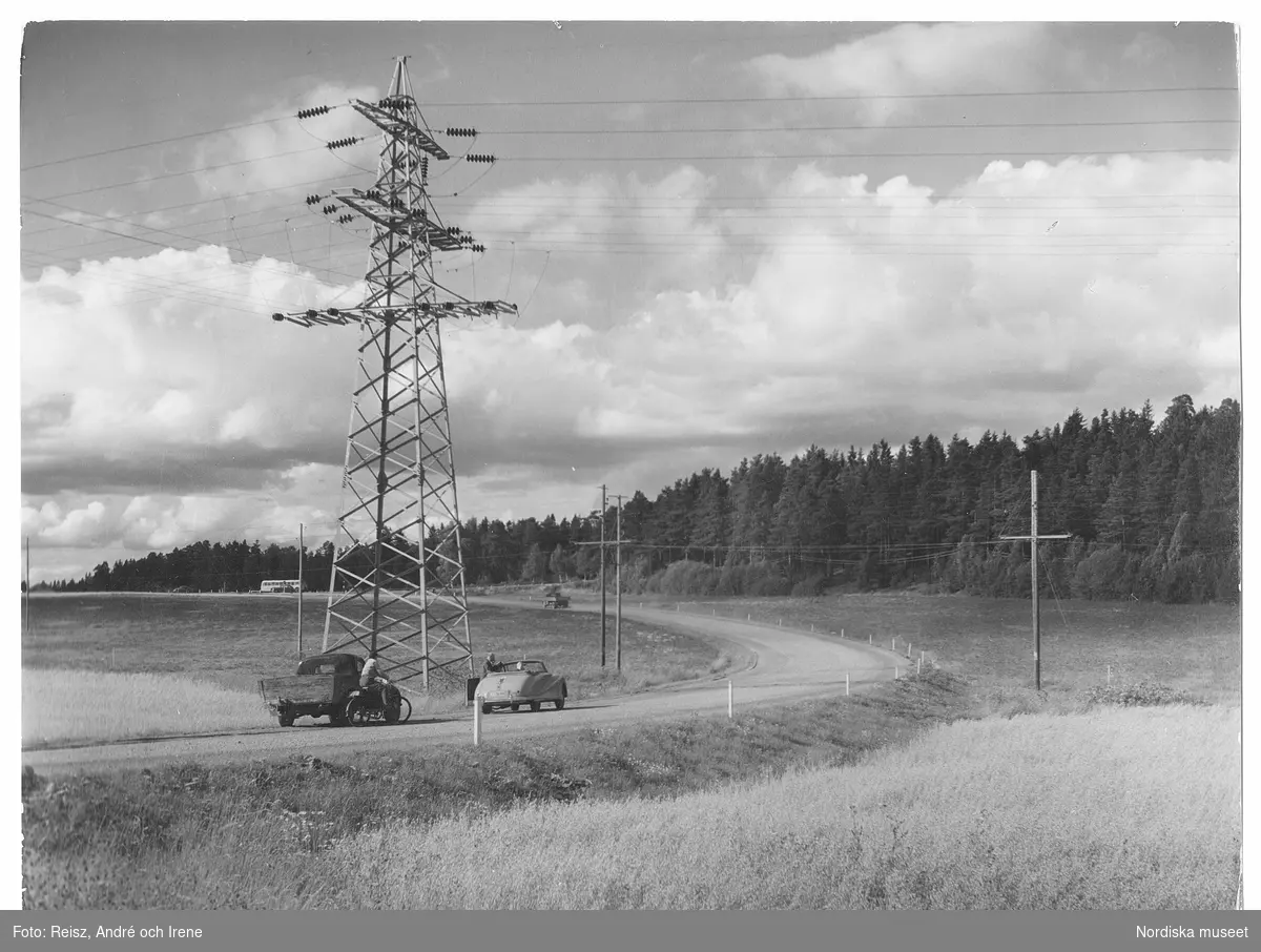Uppland. Elektrisk kraftledning i Uppland. Bild över en landsväg som slingrar genom lanskapet samt flera olika typer av fordon. En av bilarna har stannat invid vägkanten.
