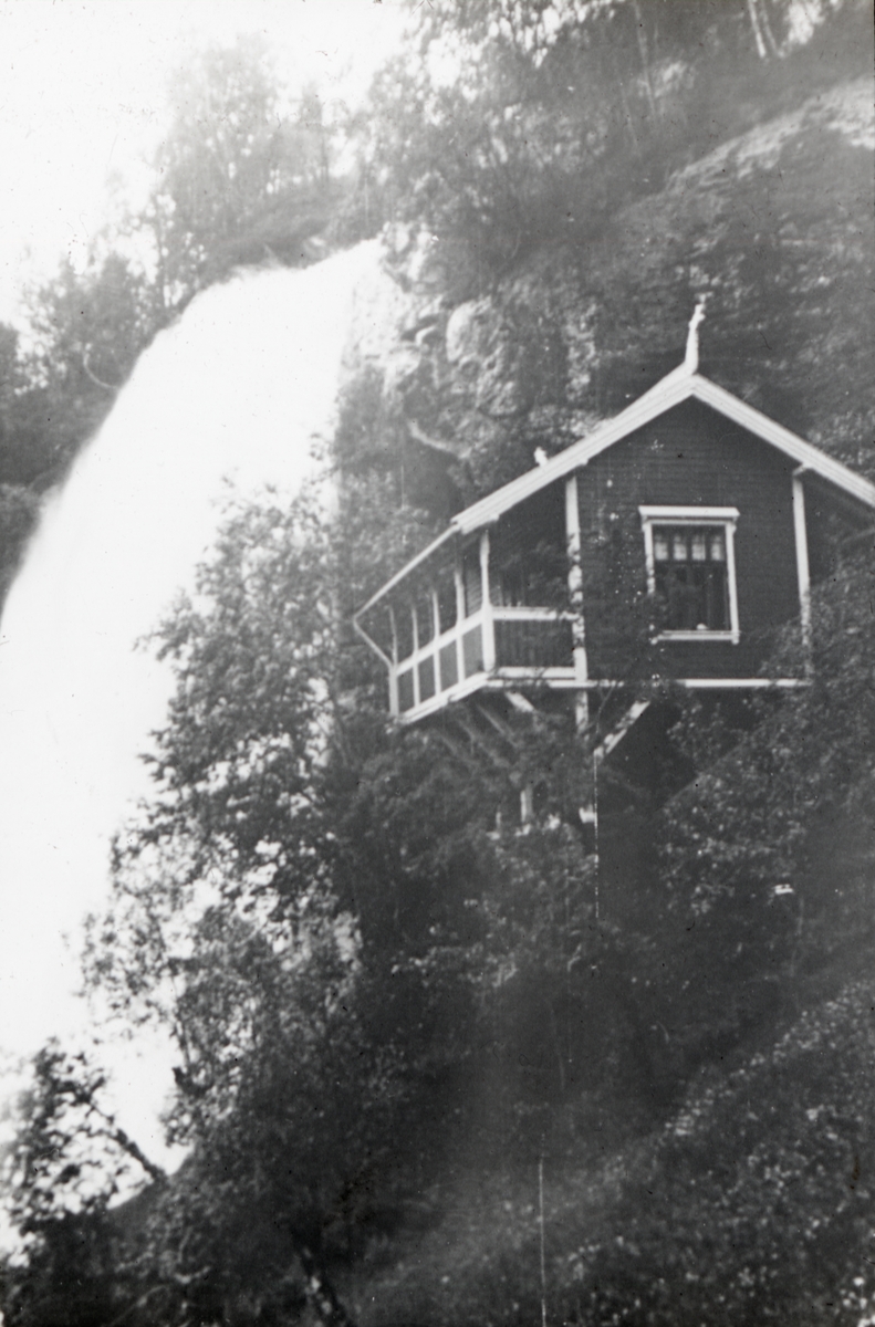 En liten hytte ligger i en bratt fjellskråning ved Steindalsfossen.   Bildes tittel stammer fra påskrift på lysbilderammen av Anders Beer Wilse.