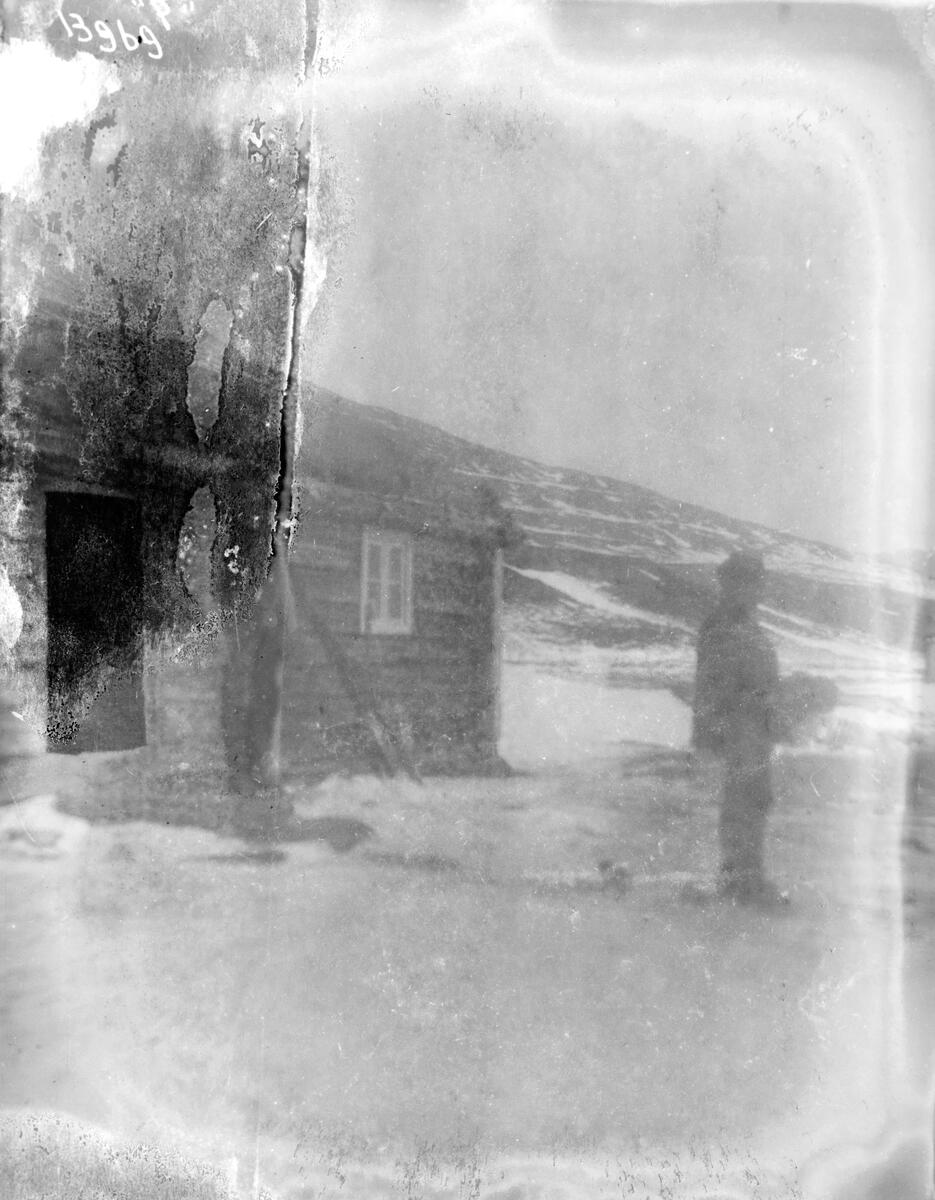 To menn står i snøen utenfor en laftet tømmerhytte eller seterhus.