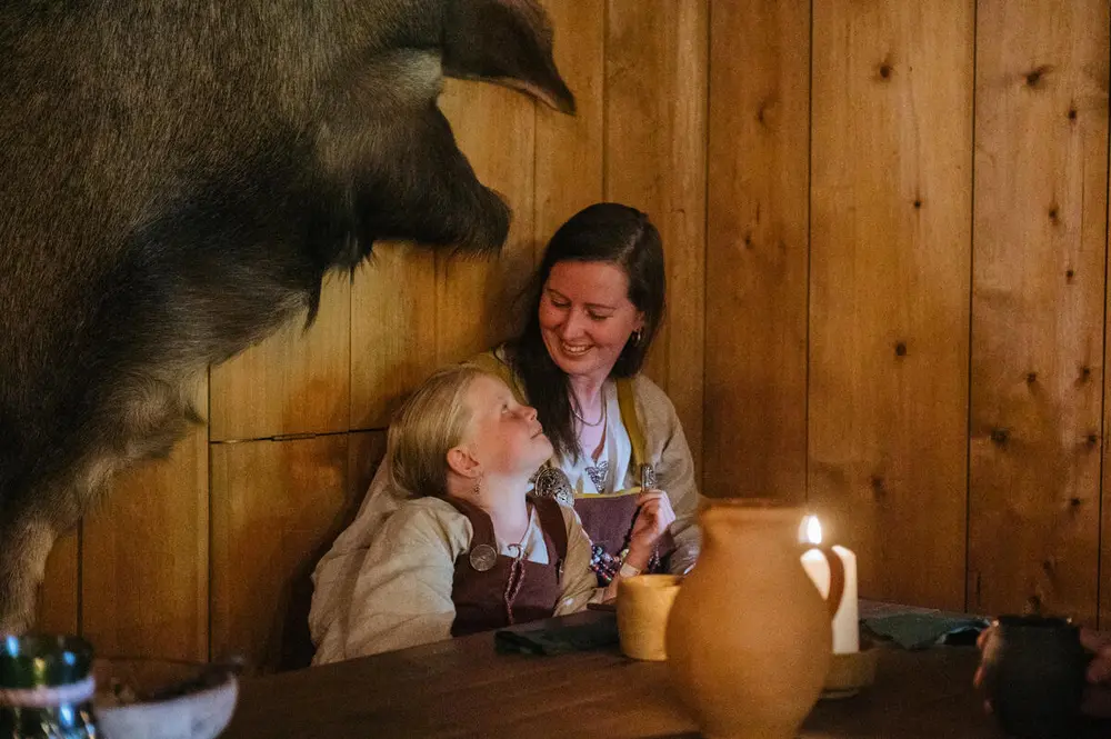 En kvinne og et barn sitter inntil en vegg. De ser på hverandre og smiler. De er ikledd vikingklær