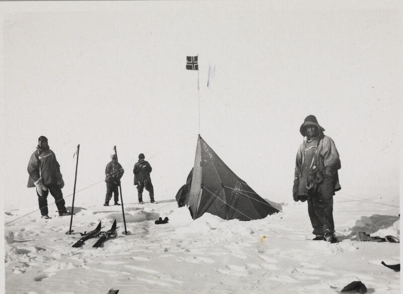 Sort hvitt bilde som viser fire menn og et telt. Det er fra Sydpolen. Mennene er godt kledd og det er mye snø på bildet