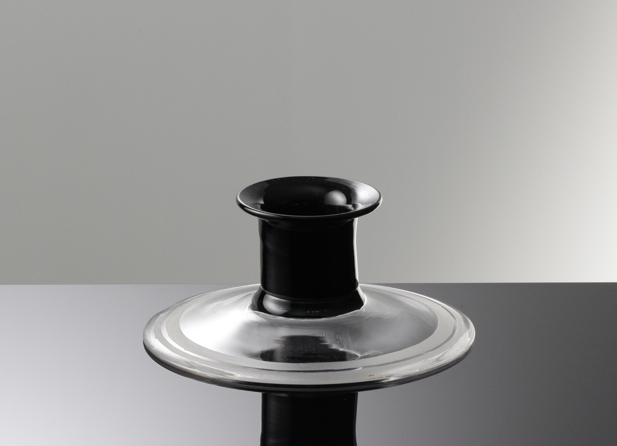 Formgiven av Simon Gate. Ljusstake med svart cylindrisk ljushållare med utsvängd mynningskant. 
Skivfot i ofärgat glas, med mattslipad randdekor i kanten.