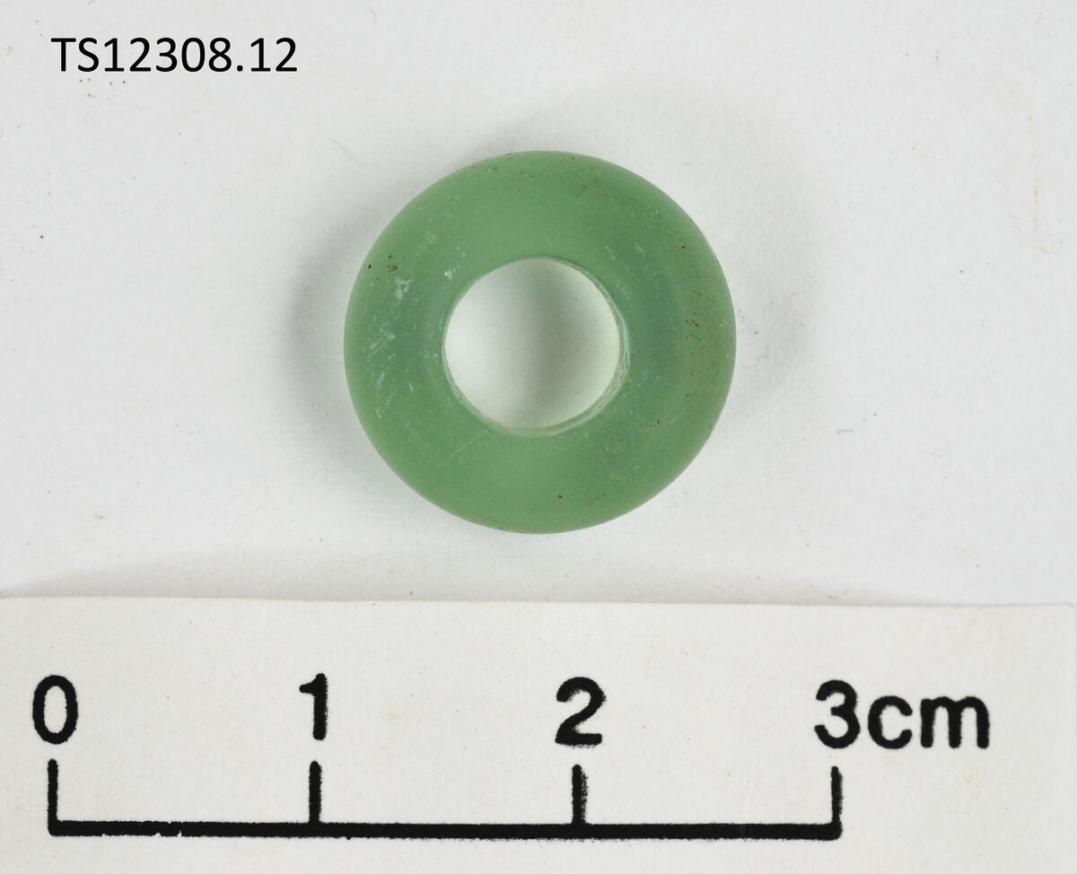 1 stk perle av glass. Lys pastellgrønn. Rundede sider og konisk, rettvegget hull. Svært homogent glass, ingen uregelmessigheter, hjull eller luftbobler - tyder p høy kvalitet. 1,5 cm i diameter, 0,6 cm tykk.