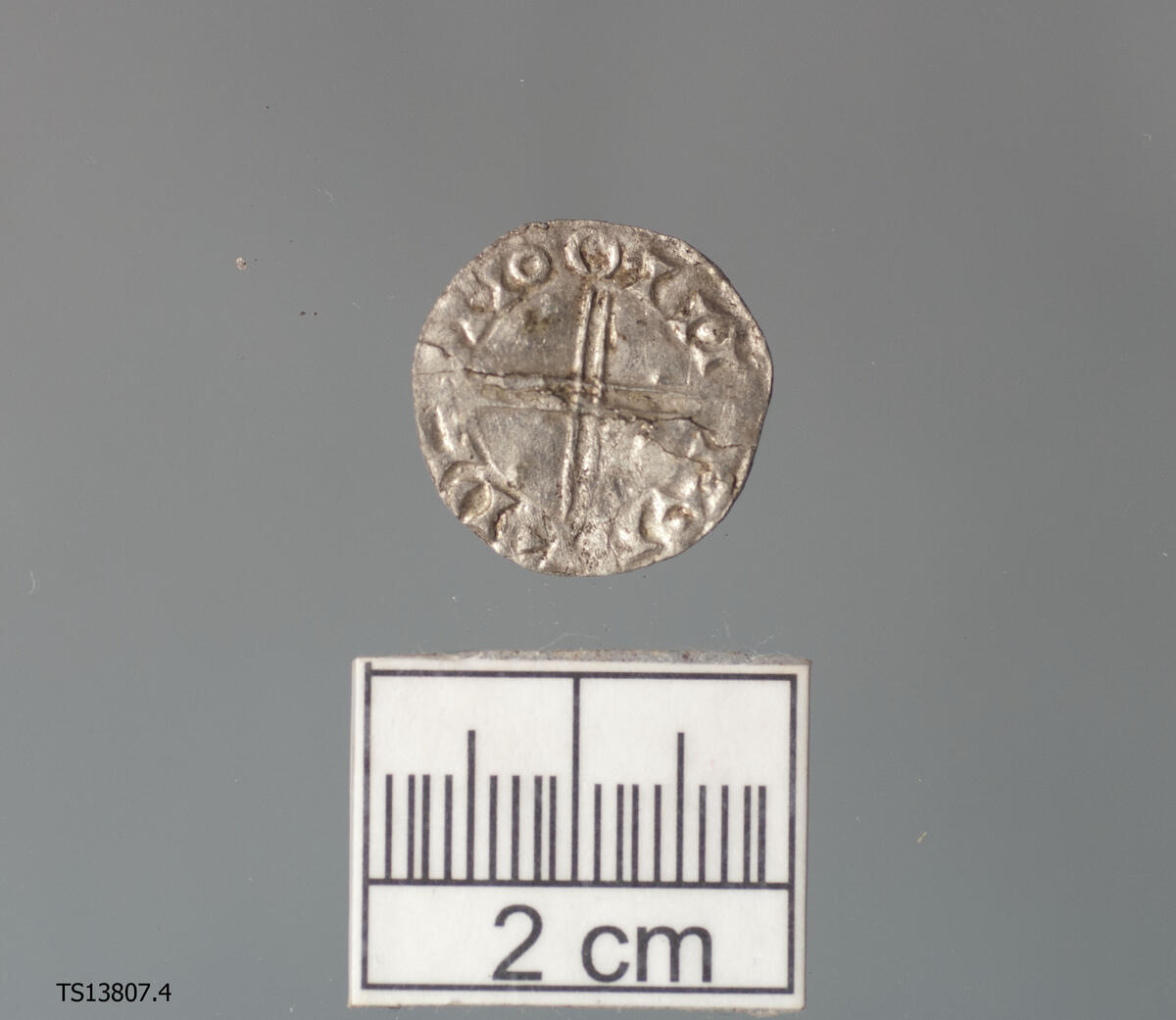 Mynt med  mannsperson i profil mot venstre som holder et septer og med uleselig skrift rundt/Dobbelkors med uleselige tegn bak. Finners forslag; Penning slått under Olav Kyrre 1067-1093. 