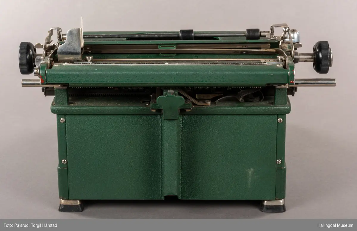 Grønn skrivemaskin av merke Halda. Den har to røde og tre gullstriper som går som en linje rundt øverste del. Noe slitasje på tastatur. 