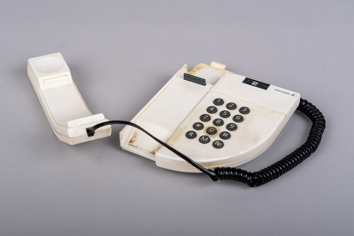 Ericsson bordtelefon.
Apparaten består av telefonrør, teelfon og spiralkabel. 
Hvit telefon med 15 svarte trykk knapper: tall (0-9), stjerne, nummertegn, funkjsonsknapper. 

Telefonen er misfarget, brune bruksmerker på telefonrør og rundt knappene, spesiell ved tallet 0.

På bunnen klistemerke med artikkel kode:
DBCN 2120700/987 R1A
ETM 9107 D. Design