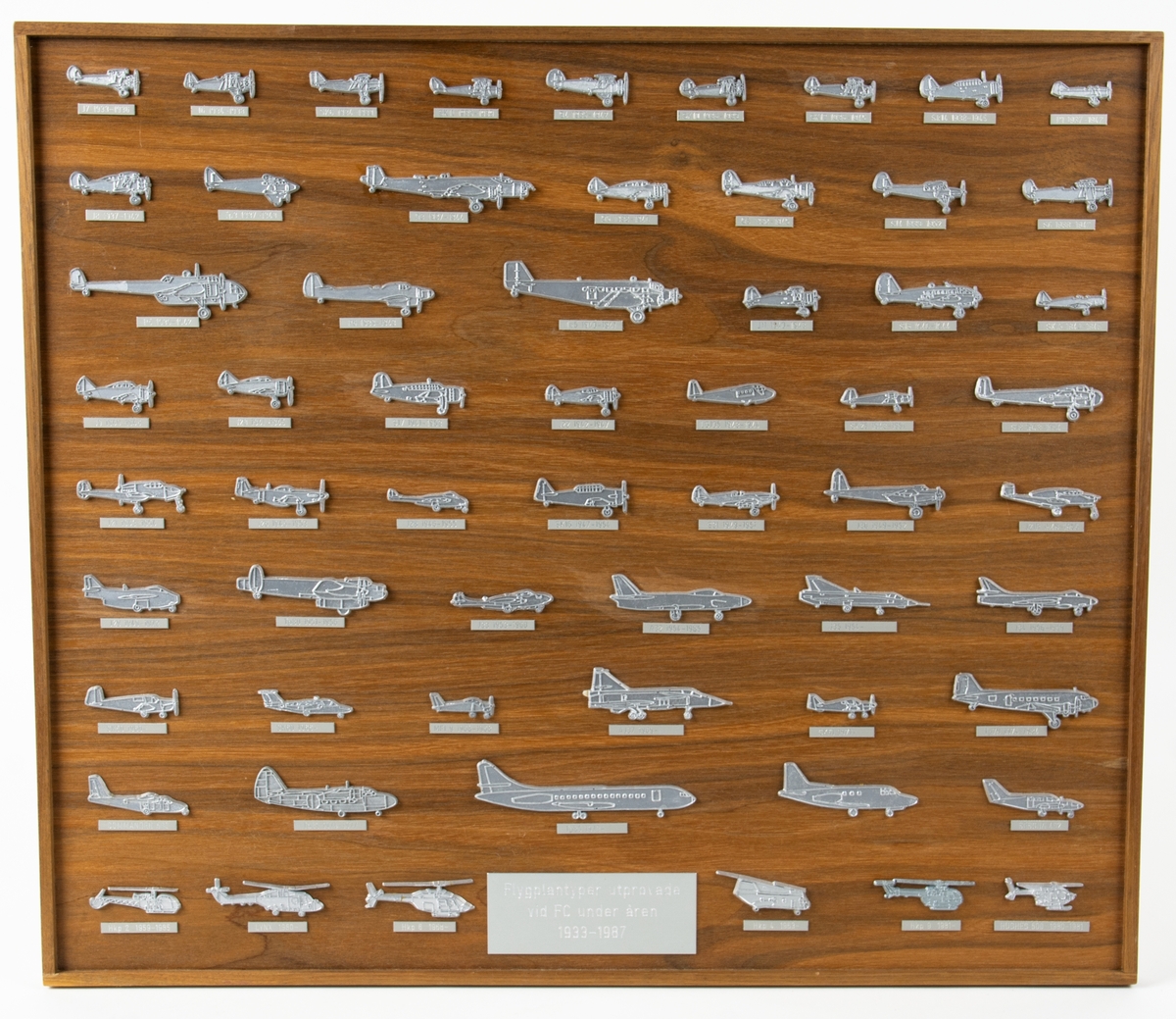Tavla med flygplanstyper utprovade vid FC under åren 1933-1987. På tavlan av trä är små silverfärgade flyglan av gjuten plast limmade.