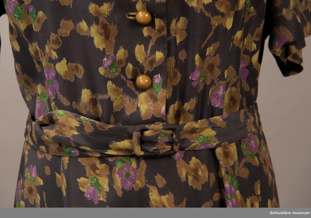 Klänning i konstsilkematerial med guld lila och grönt, blomster på lilagrå botten. Klockad kjol kort ärm och stor krage. Knäpps med tre knappar fram. Hör ihop med jacka UM.19787 i samma tyg som även syns på bild 1-3 samt 9-10.

Kjollängd 66cm.