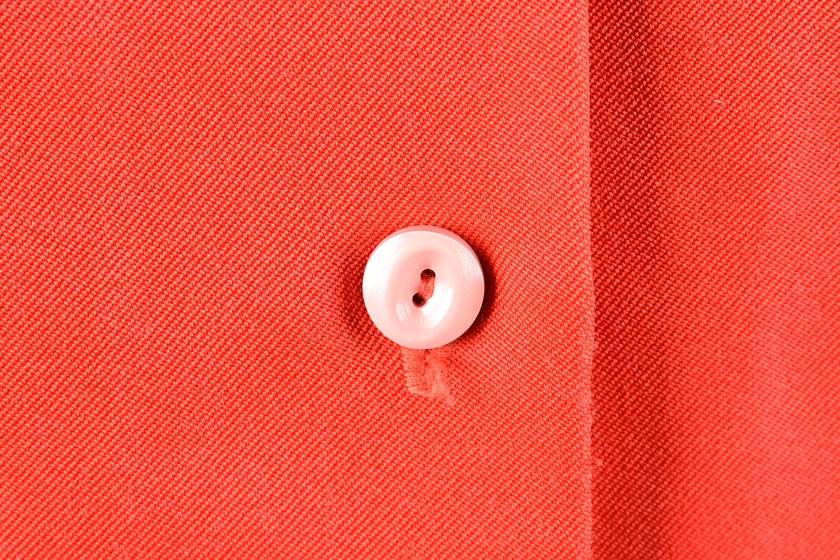 Arbeidsbluse som er kneppet i front med fem knapper samt mansjetter på ermene som lukkes med en make knapp. Blusen har en lomme på hver side av åpning i front. Den har hemper i sidene til belte. Kraven er foret med stoff som har geometriske motiv. Bærestykke og lommer er pyntet med make stoff i smale bånd.