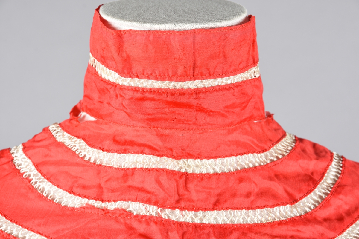 Bluse av tynn rød silke, åpen bak (hekter). Høy hals med spiler. Pyntet foran med kniplingsmotiv og hullsømsrader på armene og oventil.