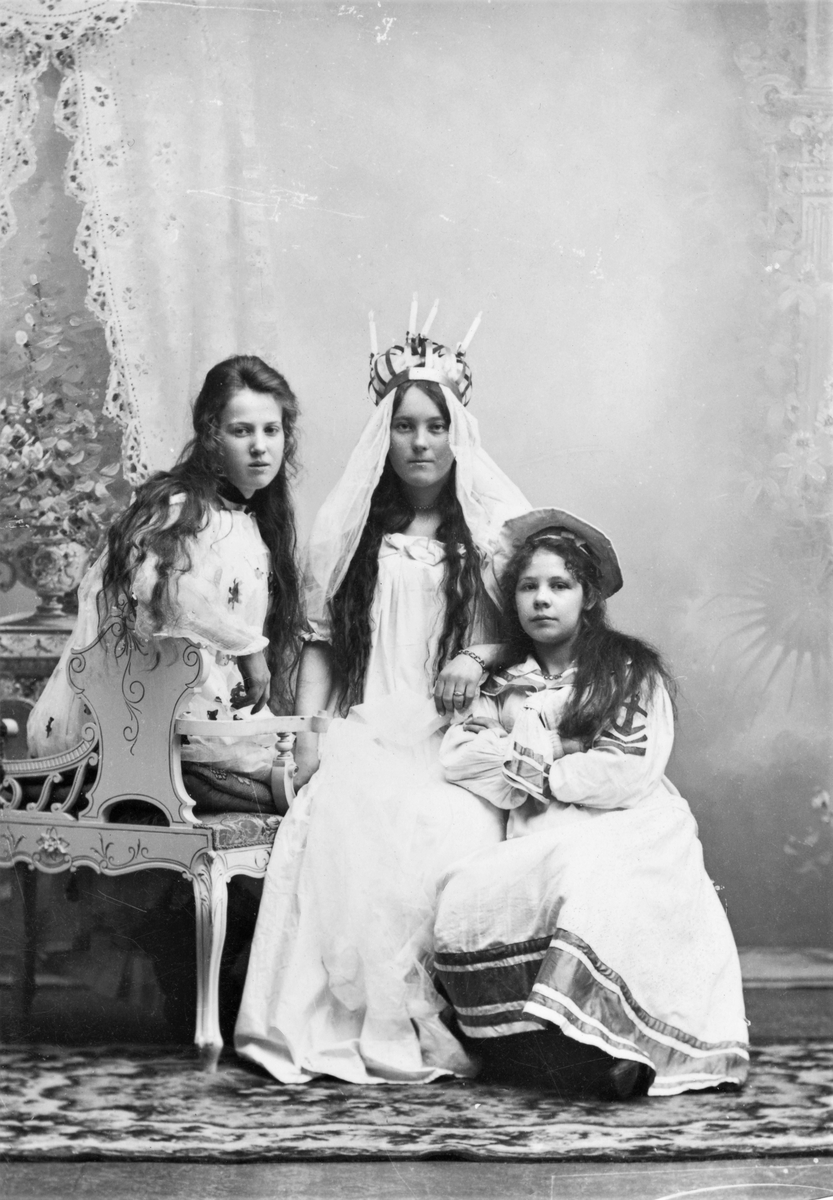 Ateljéfoto. Tre unga kvinnor flickor på en soffa, där den i mitten är klädd till Lussebrud, Lucia. Flickorna heter: Alida Carlsson, Selma Thorén och Elin Österberg.
(Eventuellt är fotot taget av Mathilda Ranch.)