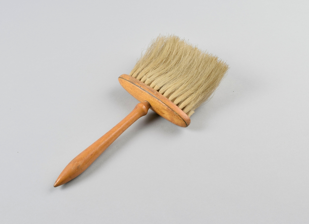 Flat børste med lange buster festet til oval plate med dreiet håndtak.
