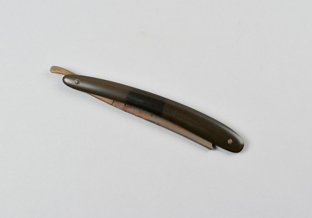 Sammenleggbar barberkniv med stålblad, skaft i svart celluloid. Bak på skaftet er det satt inn et stykke hvit celluloid. Holk av messing.