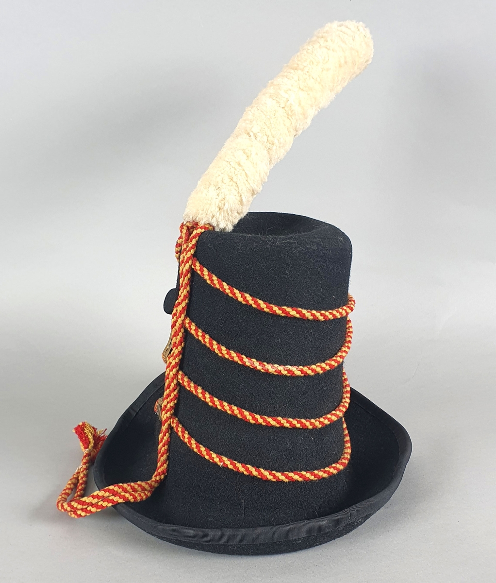 Svart hatt av filt med skinnreim på innsiden og med hvit dusk av ull på toppen og med røde og gule flettede bånd rundt pullen, festet med gullbånd og knapper. Hatten er en kopi av en offisershatt.