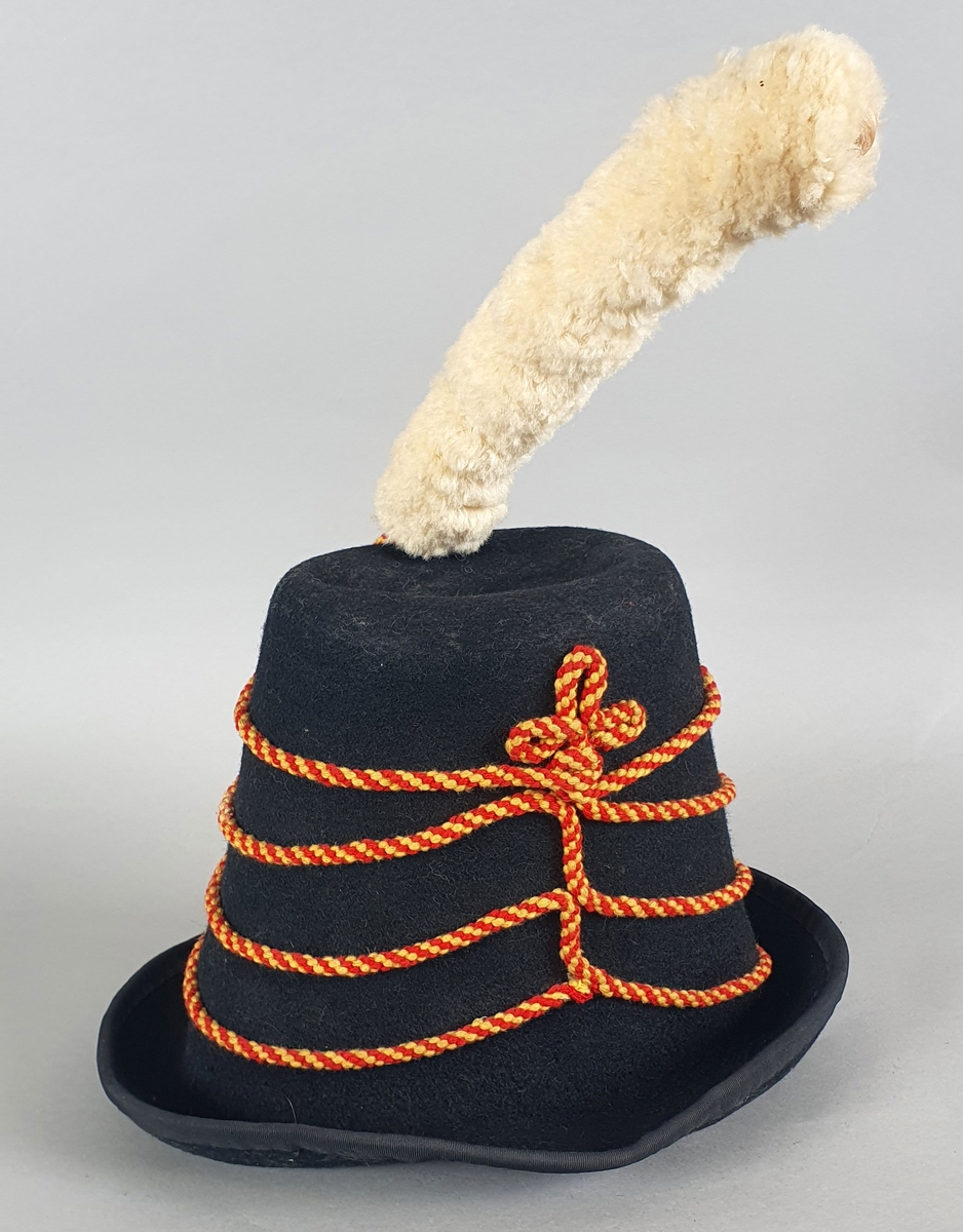 Svart hatt av filt med skinnreim på innsiden og med hvit dusk av ull på toppen og med røde og gule flettede bånd rundt pullen, festet med gullbånd og knapper. Hatten er en kopi av en offisershatt.