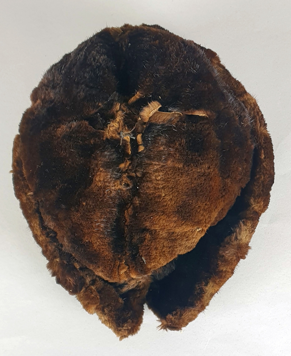 Mørkebrun hatt av pels (mink?), fôret med mørkebrun bomull. Brem som brettes opp langs pullen, med en splitt midt bak. Skinnet er revnet på flere steder.