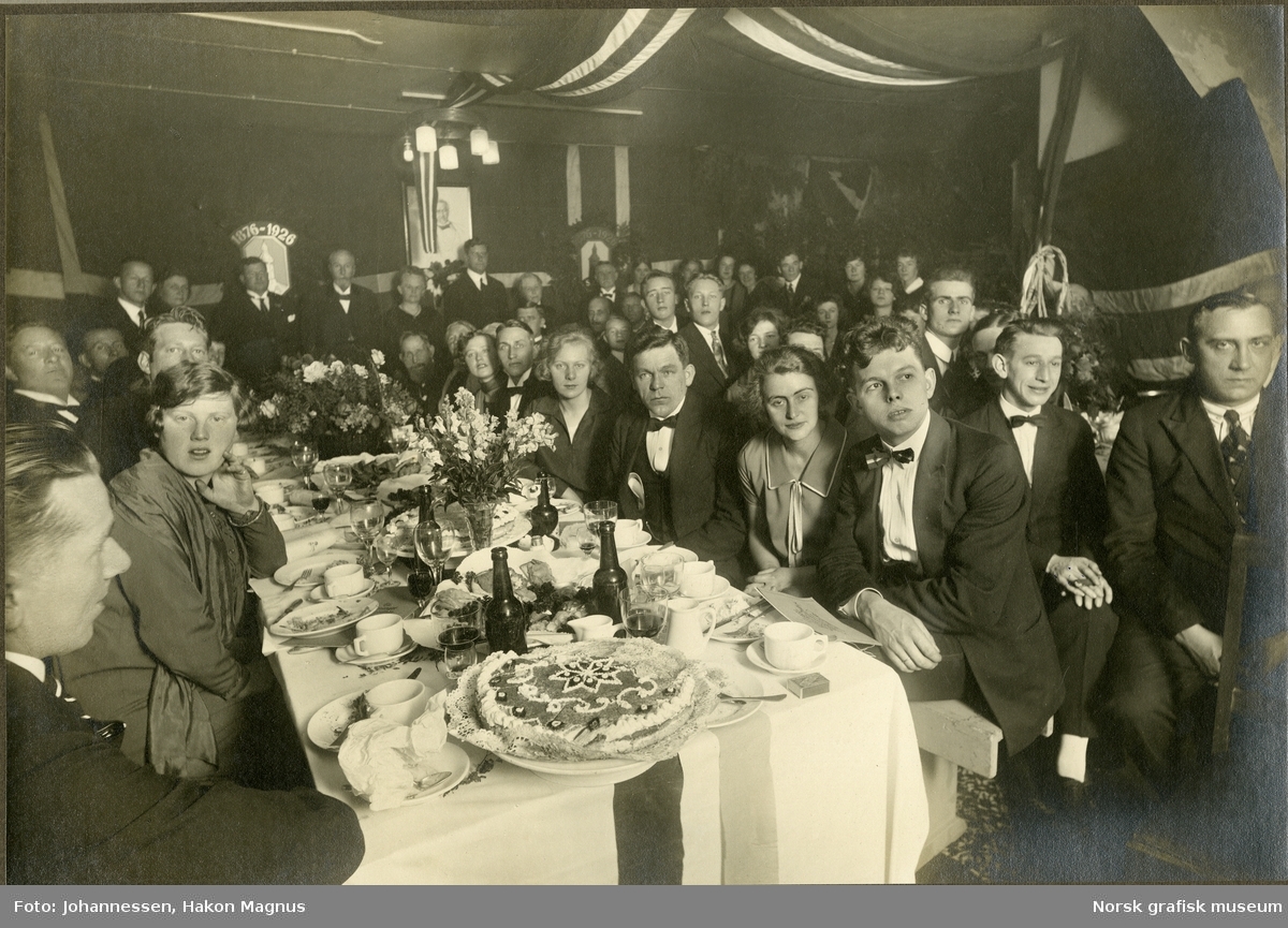 I en festpyntet sal sitter mange mennesker til bord dekket med kuverter, blomster og kake.  I bakgrunnen ser vi logoen til Stavanger Lithografisk Anstalt, sammen med tallene 1876-1926.