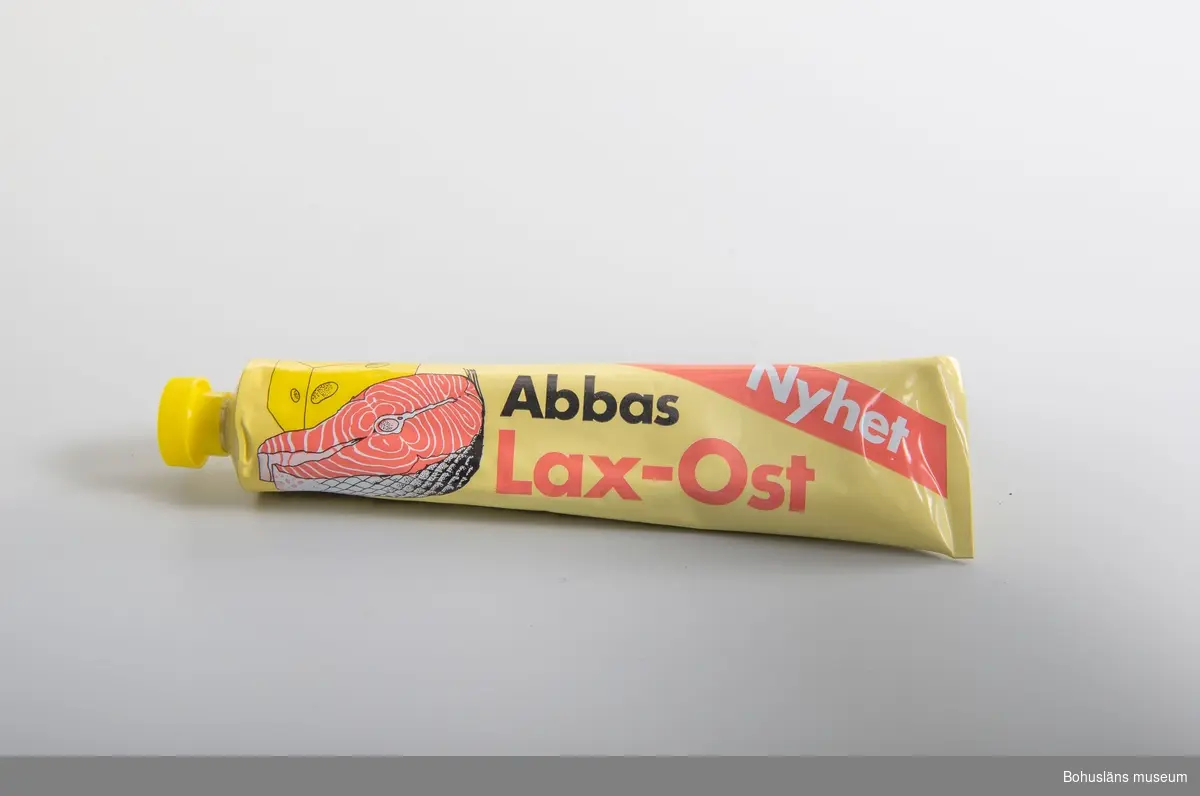 Tryckt dekor i gult,rosa,svart,vitt Gul gjuten plasthatt. Text: "Abbas Lax-Ost. Nyhet  samt produktbild  På baksidan innehållsförteckning och prismärkningskod. 
Tuben är identisk med UM026808
Om givaren se UM026667
