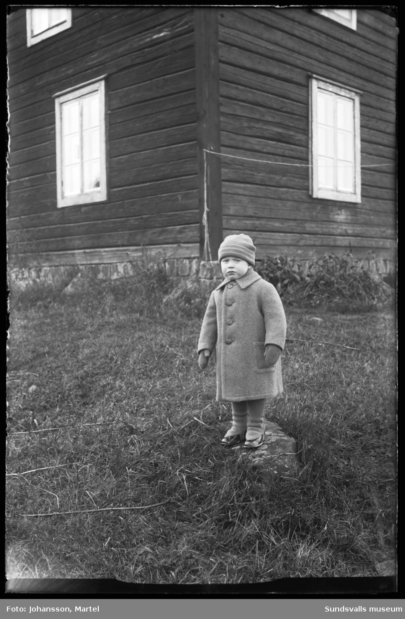 Porträtt av en flicka och pojke, troligen syskon, klädda i varma ytterkläder och stående hand i hand framför en timmerbyggnad. På bild två endast pojken.
