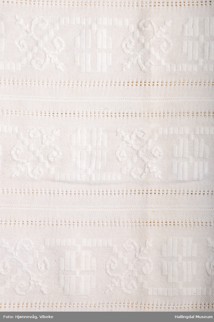 Bomullsduk vevd i toskaft, sydd sammen av 5 "remser" bomullstøy som hve er ca. 18 cm brede. Brodert med hvitt perlegarn.