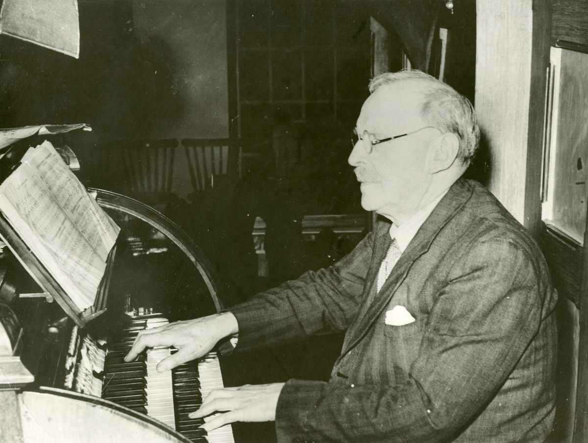 Portrett av en eldre mann som spiller på et orgel. Mannen er iført dress og slips.