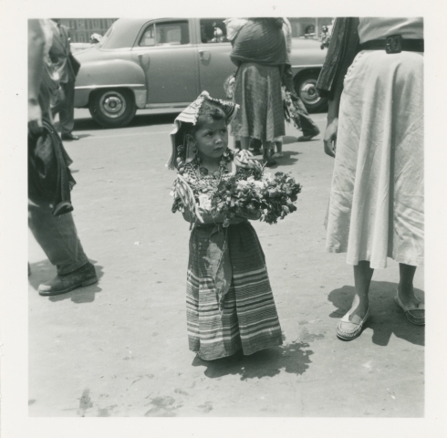 En liten jente i mønstret kjole holder en bukket blomster. Barneparaden, Mexico.