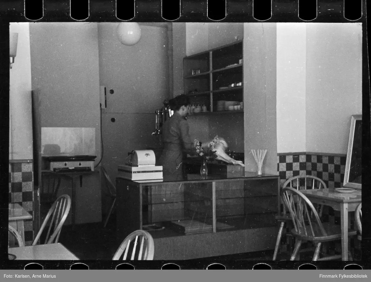 Foto av interiør av kafé i Kirkenes, ukjent sted og ukjent kvinne bak disken

Foto antagelig tatt på slutten av 1940-tallet, tidlig 1950-tallet 