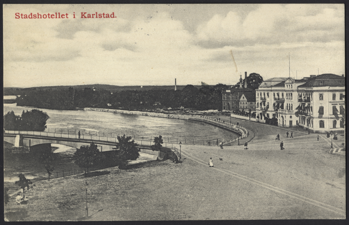 Vykorten föreställer en vy strandkanten i Karlstad med stadshotellet till höger och en bro till vänster. Trafiken består uteslutande av gångtrafikanter.