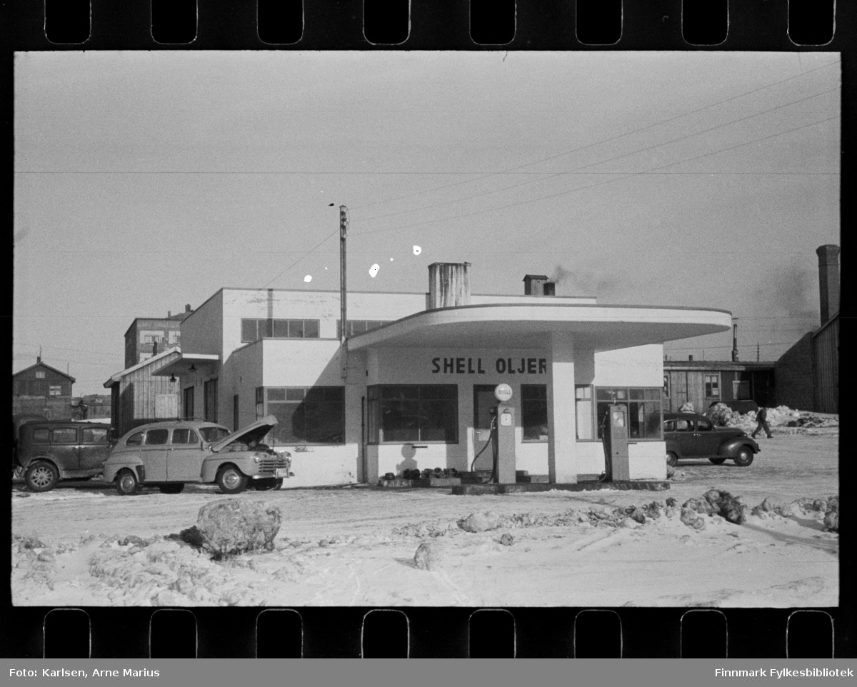 Foto av Shell Oljer bensinstasjon i Kirkenes 

I foto kan man se at gaten er en grusvei. Ved siden av bensinstasjonen ser man flere biler parkert og foran stasjonen er bensinpumper. Lys bil med panseret oppe er en Ford 1946-modell, reg.nr. Y-3300 (?) med undertekst DROSJE. Bak den en ca. 1930-modell.