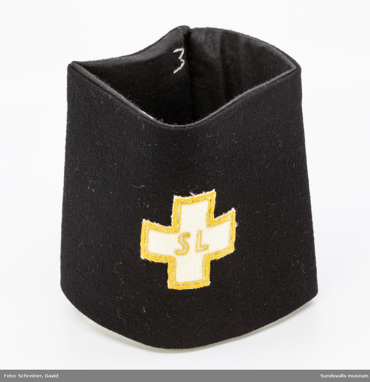 En svart armbindel som bär ett vitt kors med guldkolorerade bokstäver samt kantdekoration.