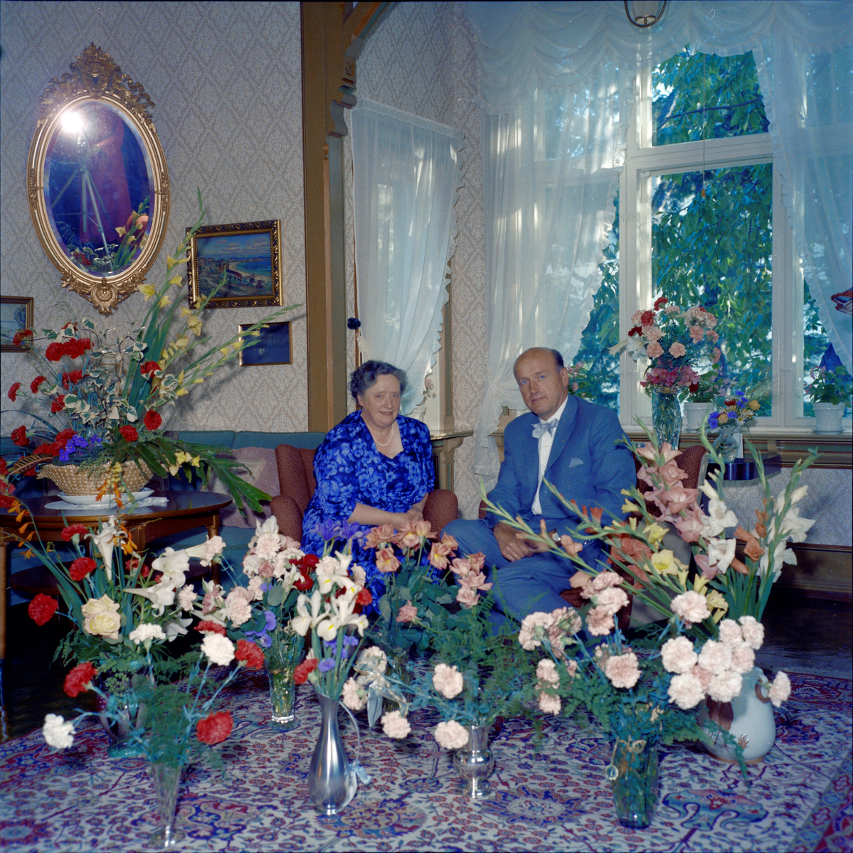 Eilif og Lilly Schrøder omgitt av blomster i anledning deres sølvbryllup. Fotografen, Rakel Schrøder, vises delvis i speilet.