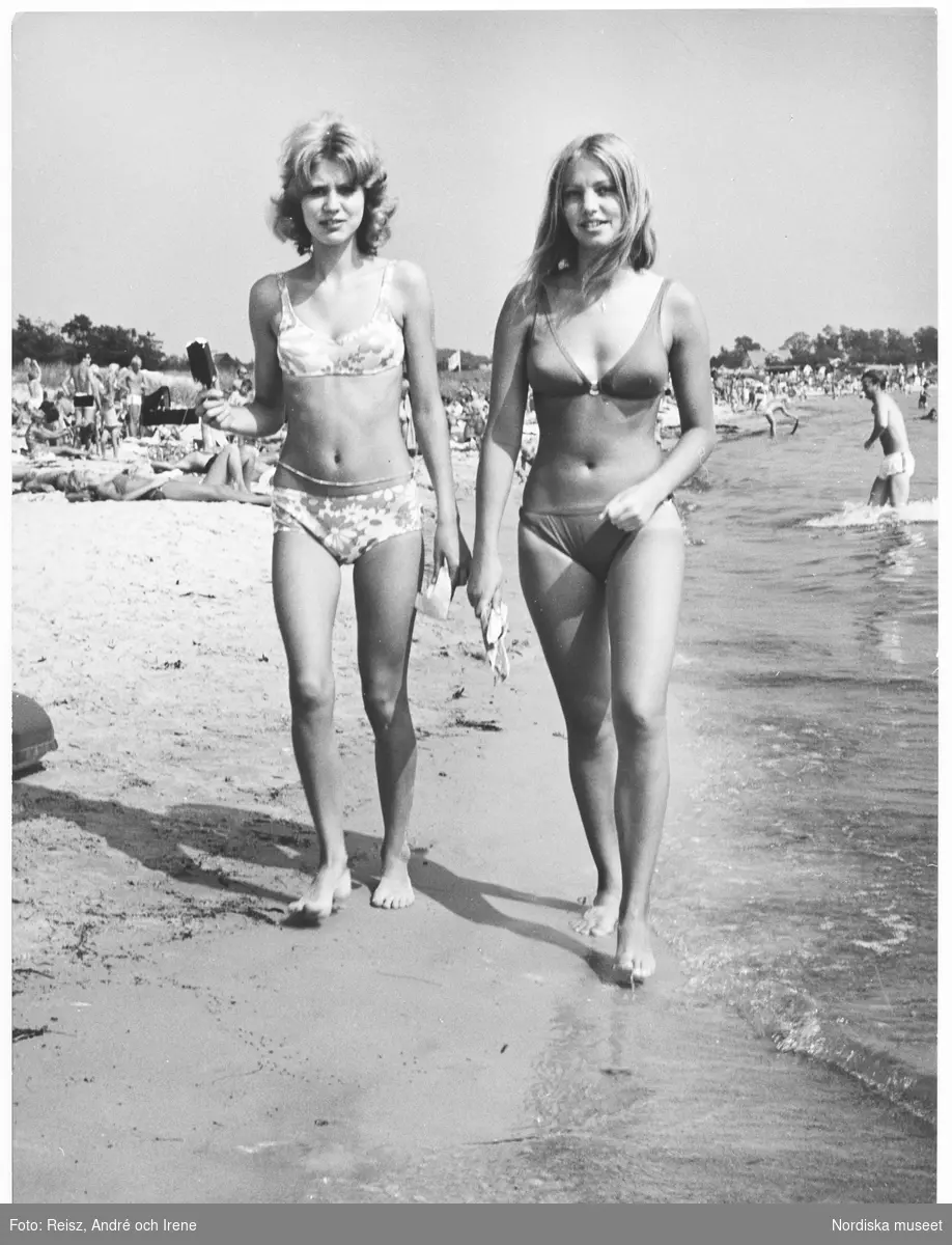 Två unga kvinnor promenerar längs den välbesökta stranden. Kvinnan till vänster håller en pinnglass i handen.