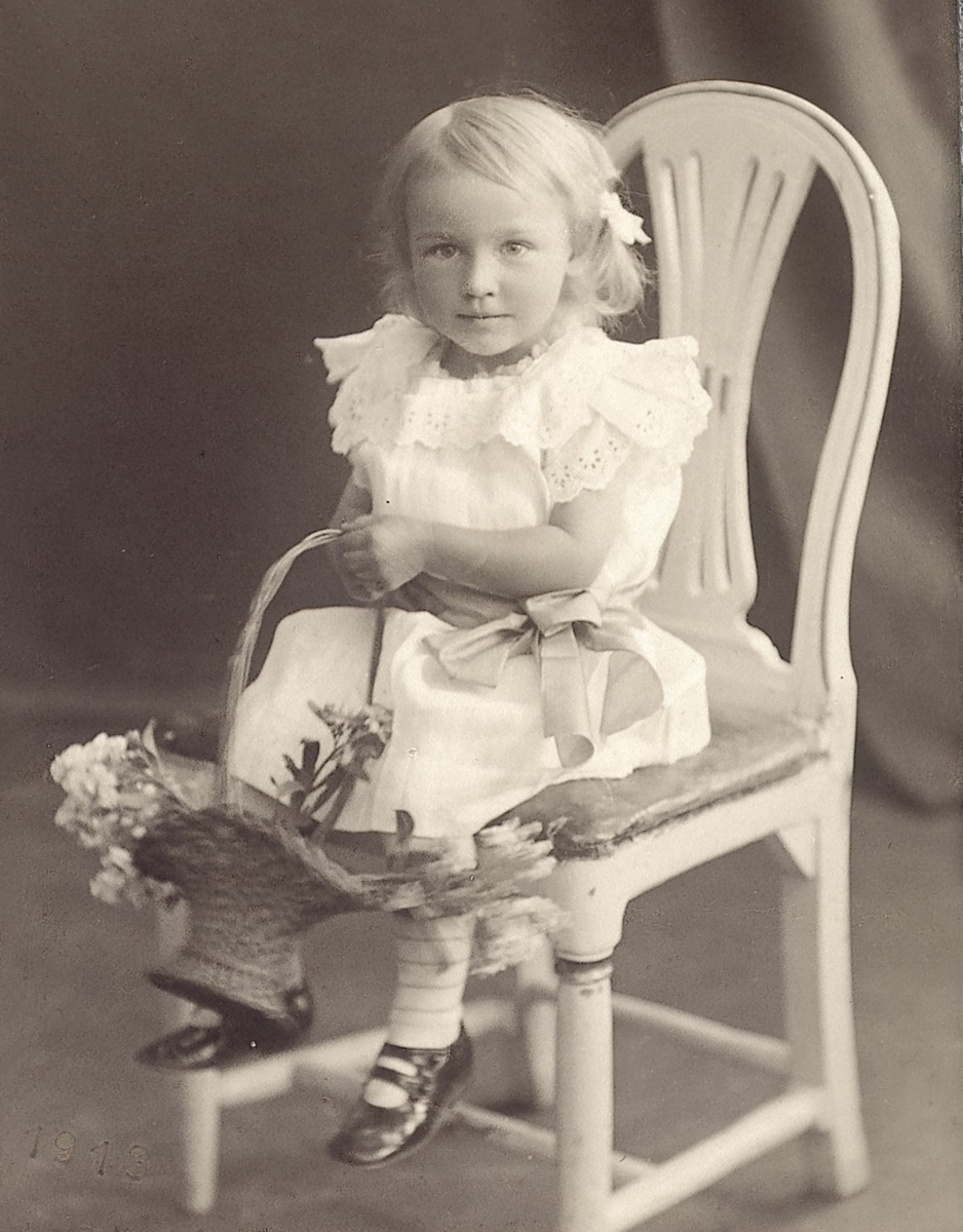 Ateljéfotografi av en liten flicka, sittandes på en stol med en blomsterkorg i händerna.