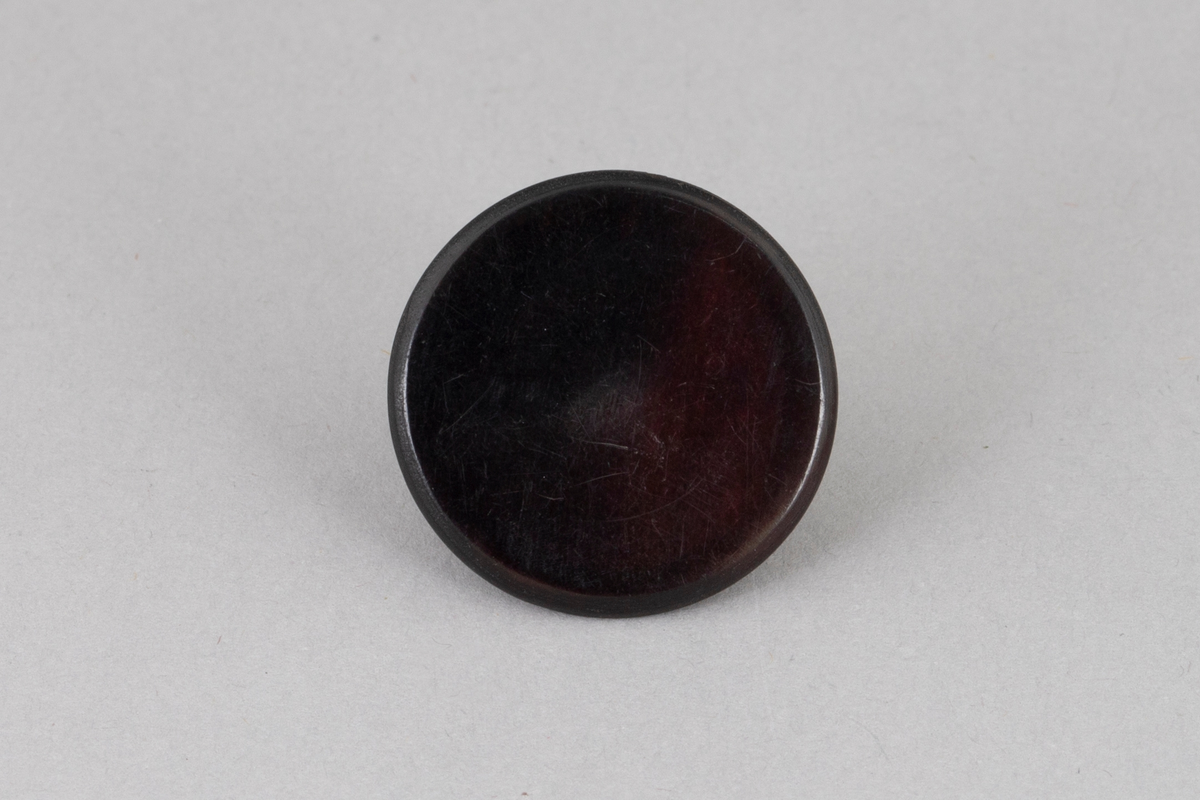 Rund svart knapp i plastlignende materiale med bakside av metall.