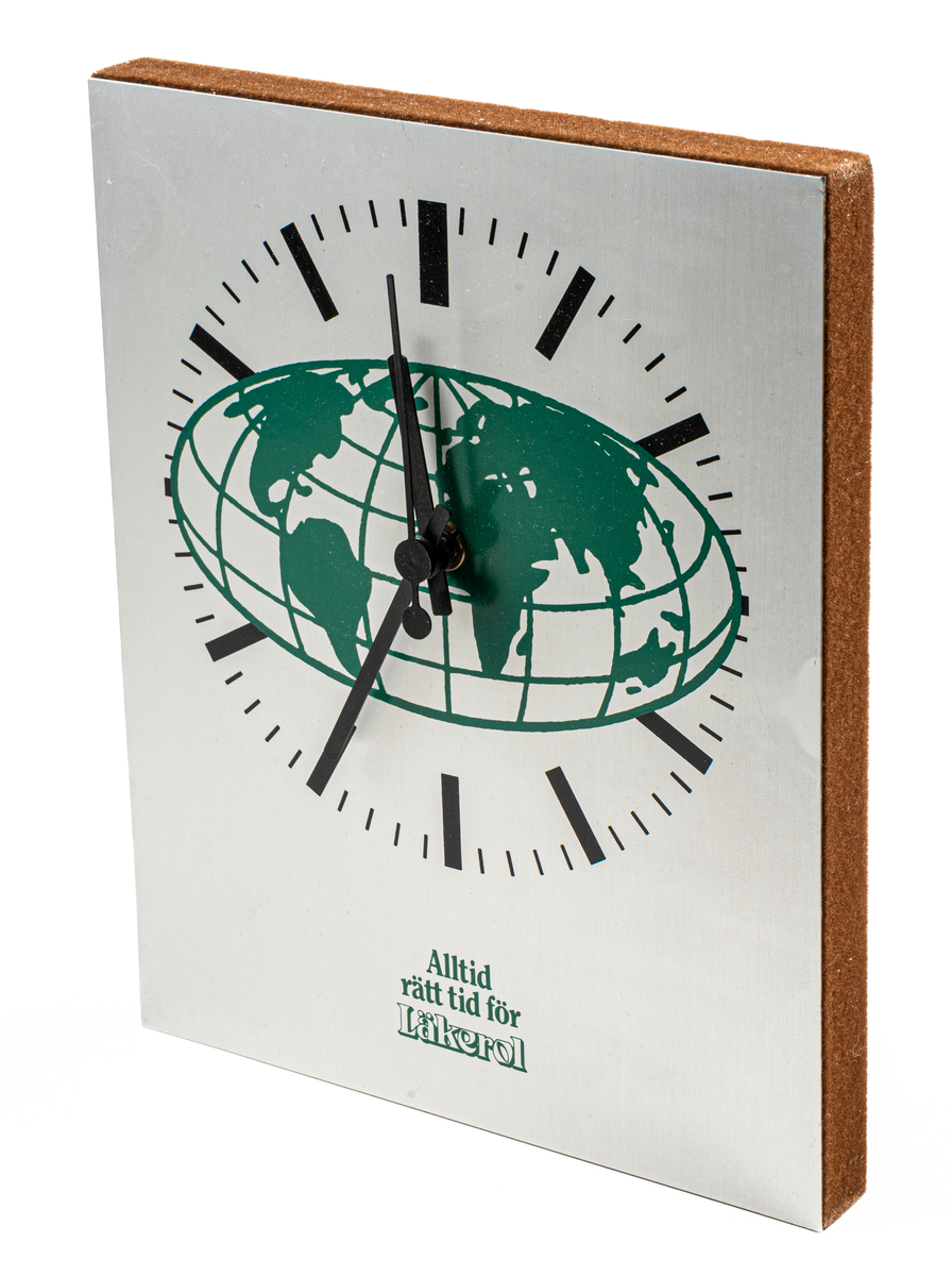 Väggklocka i silverfärgad metall, svart visare och streck för siffror, grön världskarta på urtavlan. Reklamtext i grönt: Alltid rätt tid för Läkerol. I pappkartong.