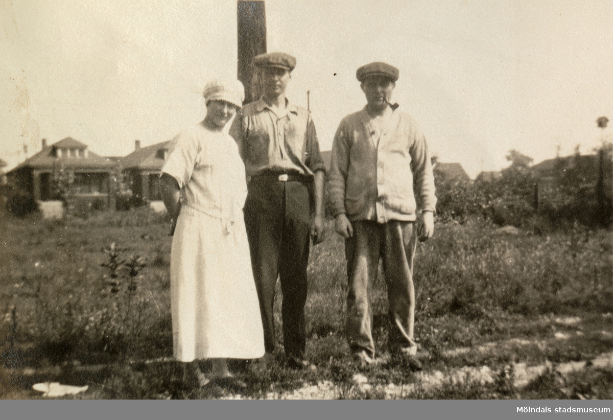 Tre personer står på en åker, troligtvis i Alaska 1924. En äng och en byggnad ses i bakgrunden. Personerna är anhöriga till givaren.