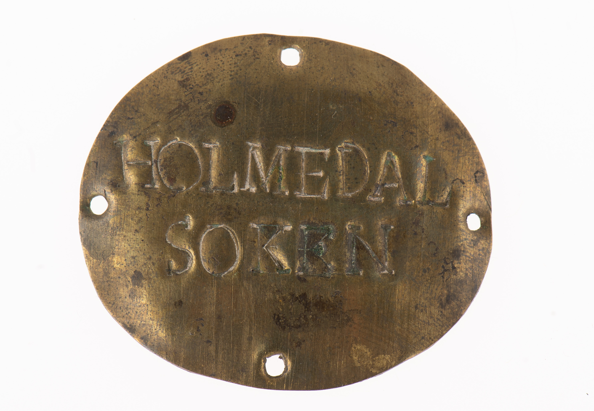 Oval mässingsbricka med fyra hål för att fästa i kläderna med sytråd. Holmedal socken.