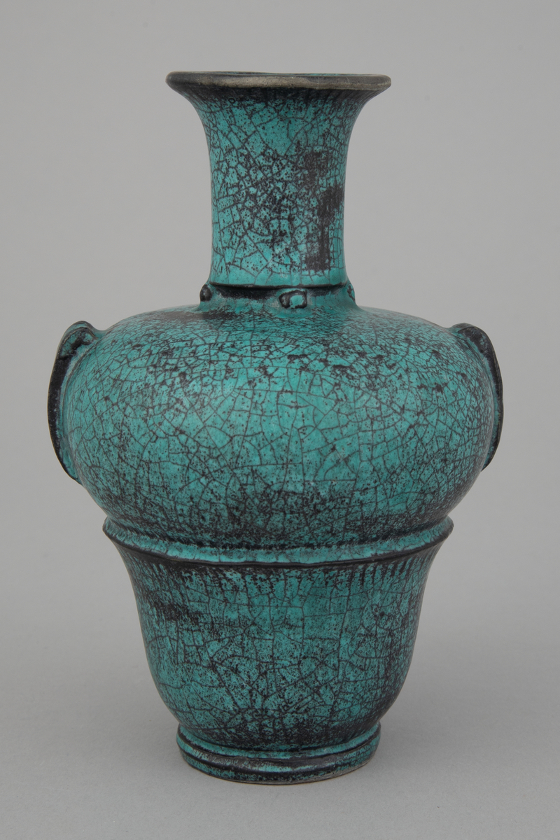 Liten vase med brede skuldre og smal hals. Vasen smalner inn ned mot foten. Den er glasert med grønn askeglasur med svarte partier, halvmatt og krakelert.