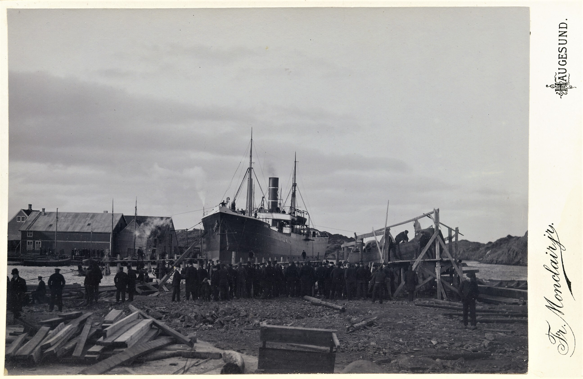 Menn og barn ser på B. Stolt-Nielsens dampskip "D/S Karmø" ved et verft. Det kan være ferdig reparert etter kjøpet i 1898. En mindre båt er under konstruksjon på land.
