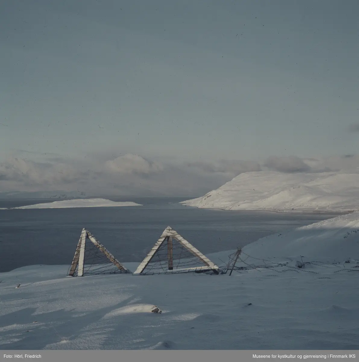 Utsikt over Sørøysundet og Melkøya i vinteren 1957/1958 fra fjellet sør for Hammerfest. I forgrunnen ser man rester etter installasjoner med piggtråd. Landskapet er dekket med snø. Bildet er muligens tatt fra Rypefjellet.