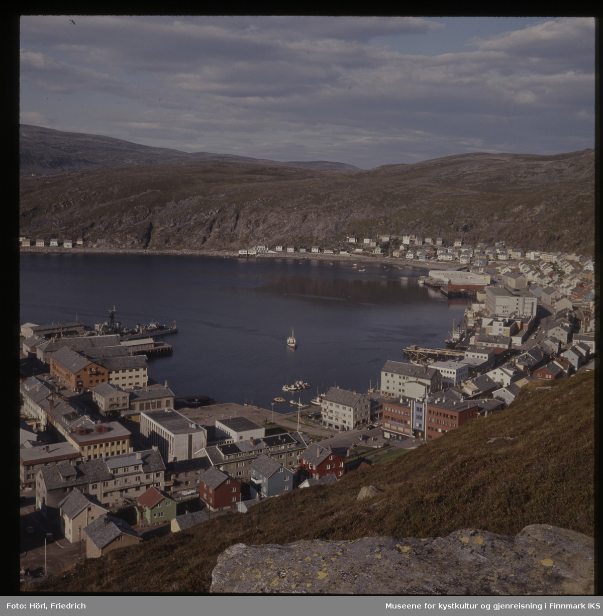 Det er sommer i Hammerfest og fra Salenfjellet ser man utover byens sentrum med rådhus, bankbygg og Findusfabrikken helt bort til Hauans materialandel ved munningen til Storelva og bydelen Molla bak den. Byen er gjenreist og fargerike hus med skifertak preger bybildet. I havna liger det ulike typer båter. Marinefartøyet på bildet er en norsk fregatt, mest sannsynlig KNM Tromsø eller KNM Haugesund.